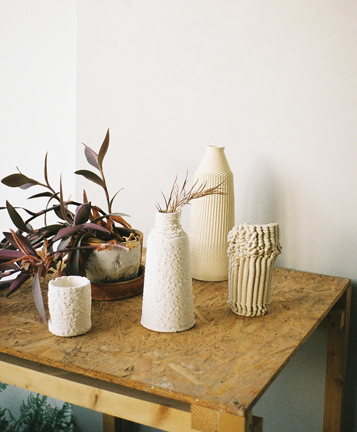 White 3D-printed vases on a wooden table, with botanical decorations. The vases are textured, with organic bumps and ridges formed through the printing process.