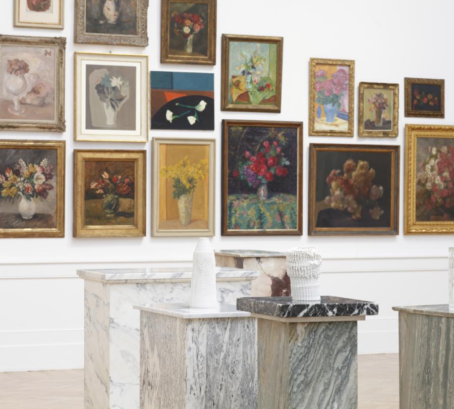 Two 3D-printed ceramic vases, each on its own pedestal, in front of a wall covered in framed oil paintings of flower vases.