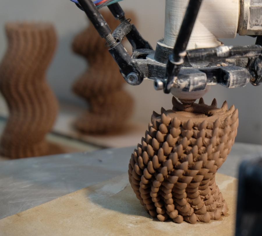 A 3D printer nozzle is in the process of extruding clay. The resulting tubular shape has a texture with multiple spike-like protrusions.