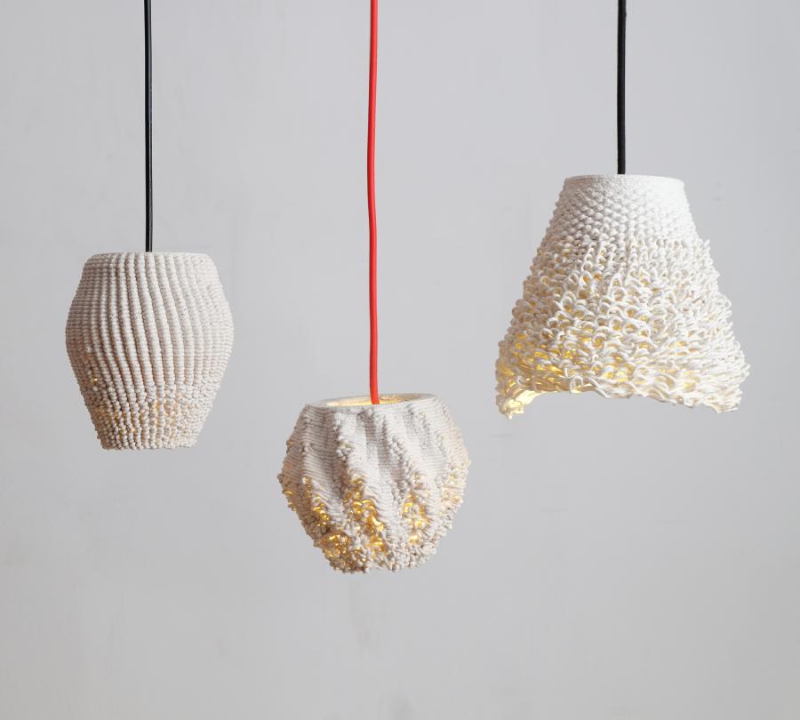 3 white 3D-printed lamps hang from thick black or red wires at different heights. They have textures similar to frayed textiles. Their light shines through the spaces between the ceramic 'threads'.