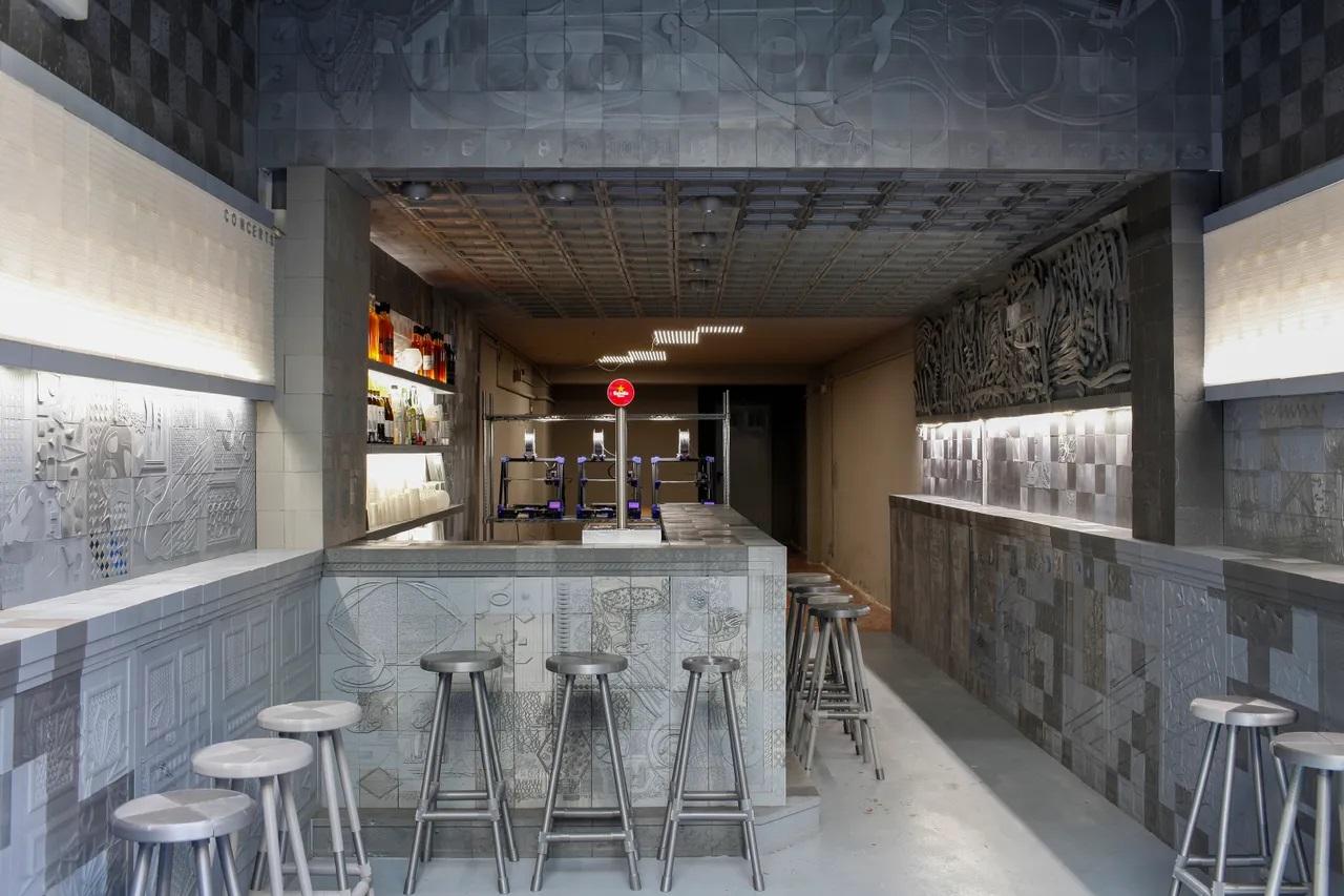 The softly lit interior of the Ex-Designer bar. The chairs and tiles covering the walls and bar are made out of 3D-printed plastic, in slighly different grey tones. The reliefs of the tiles create large scene illustrations and abstract motifs.