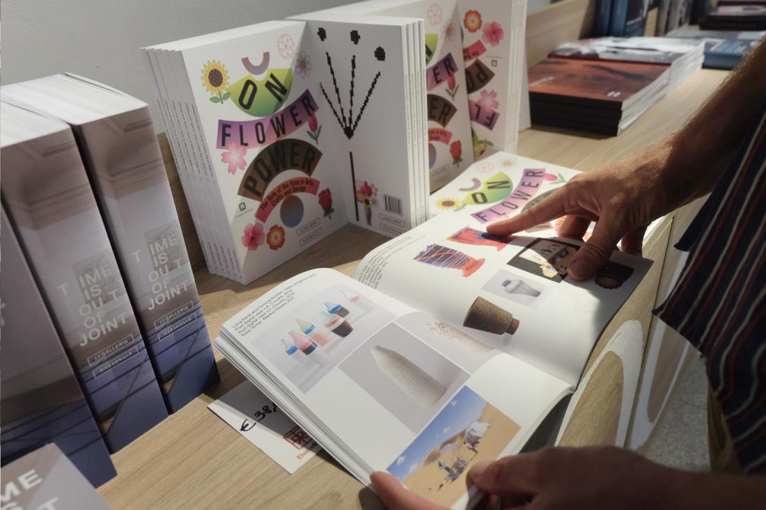 The hands of a person browsing the exhibition catalog, featuring Coudre's pieces among other pictures.