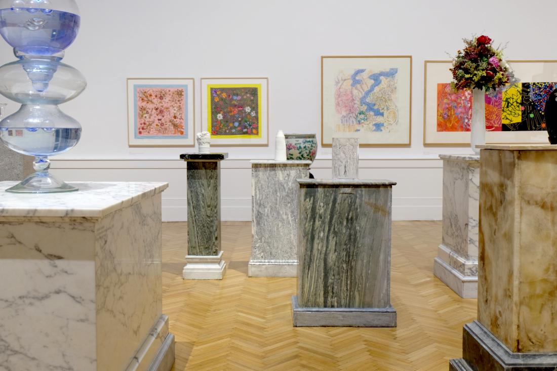 A shot of the exhibition gallery. Multiple marble pedestals with the exhibited vases on top, by different artists. There's framed paintings in the wall behind.