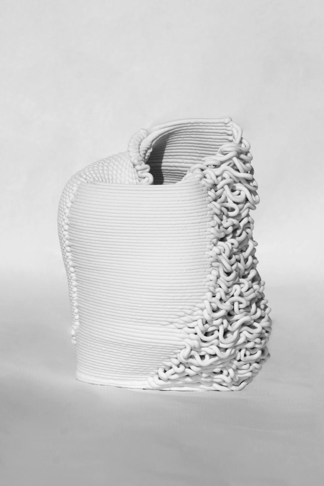 Third piece, made of printed porcelain. It has a cup-like shape, but the left upper wall is collapsed inwards. The printed layers are very loose on the right side, resembling threads. The left side has a knitted-like texture. In between these sides, it has the default 3D-print layer texture.