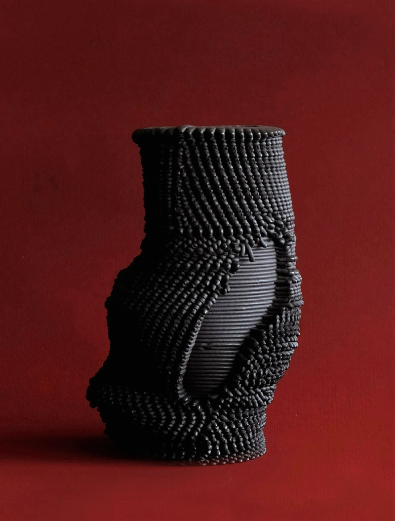 A black 3D printed stoneware vase with a tubular, organic shape. Its walls are thick and with a spiky texture, made out of printed extrusions. A patch on the piece is free from this spiky texture, showing only the printed layers.
