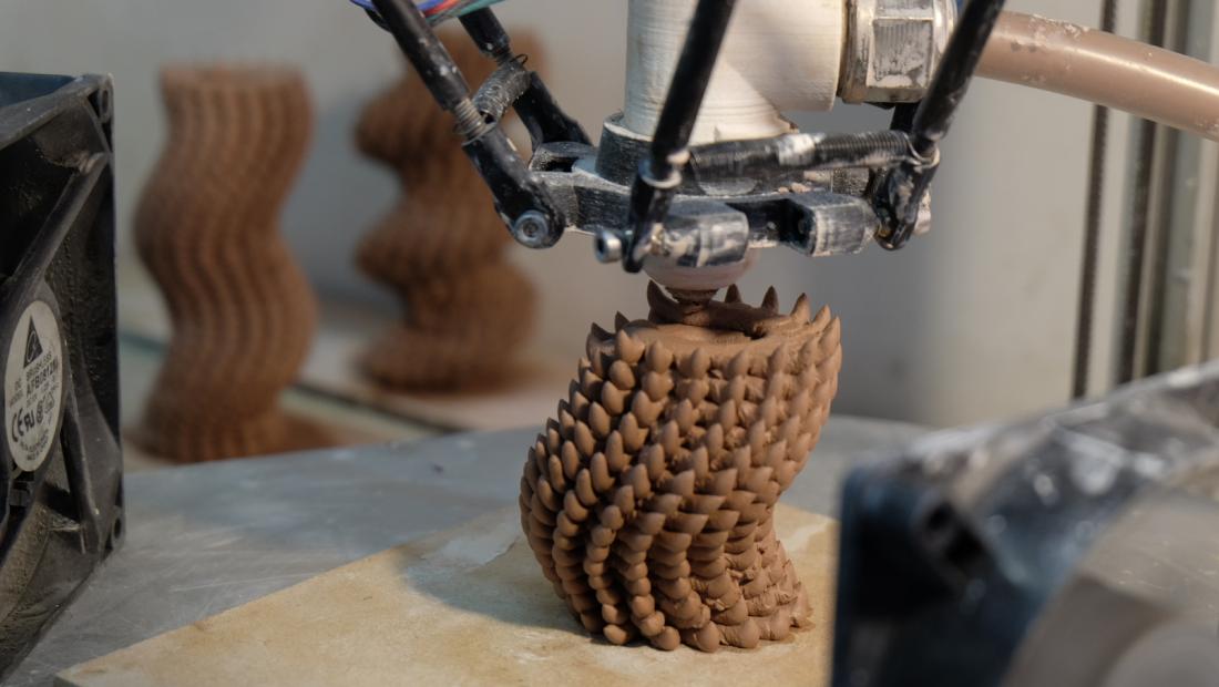 A 3D printer nozzle is in the process of extruding clay. The resulting tubular shape has a texture with multiple spike-like protrusions.