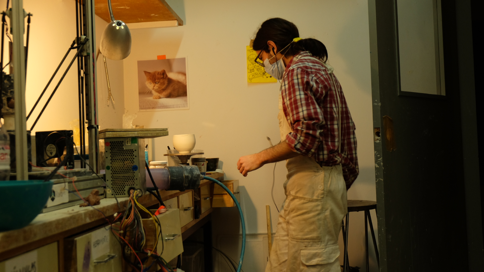 The workshop, full of 3D printers and open drawers, a picture of a kitten hung up on the wall, and Jude working.