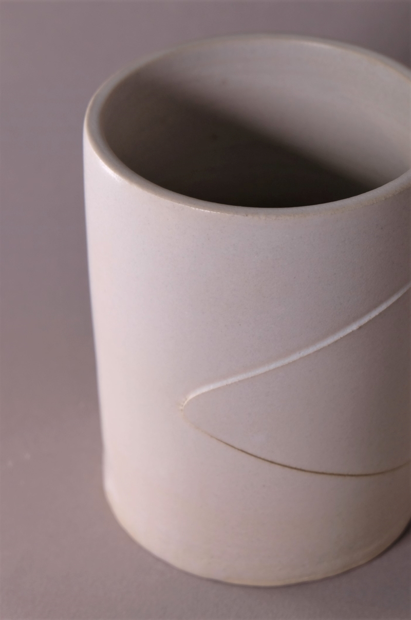 A white glazed matte ceramic piece. It is plain and cylindrical, except for a minimal triangle-shaped cut going through it, from its right side to the middle.
